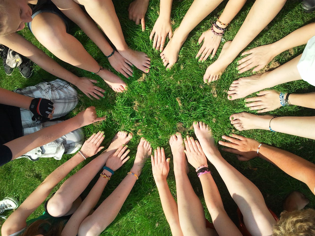 Group of friends forming a circle with their hands and feet on grass, symbolizing unity and friendship.